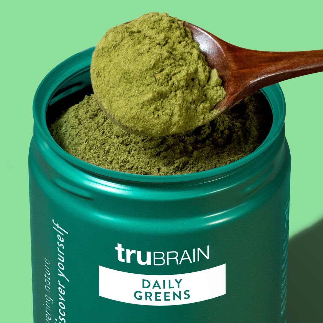 Wooden spoon scooping vibrant green powdered daily greens supplement from a teal container, highlighting the nutrient-rich product against a green background.