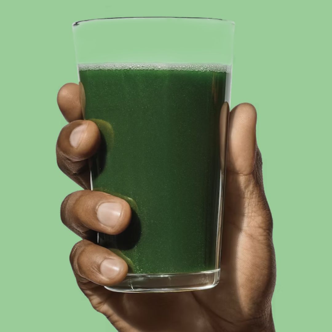 Hand holding a glass filled with vibrant green daily greens drink against a light green background, showcasing a nutrient-packed beverage.