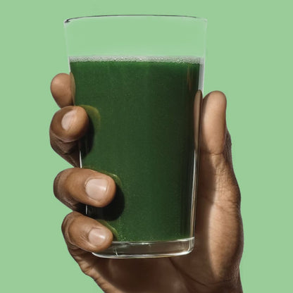 Hand holding a glass filled with vibrant green daily greens drink against a light green background, showcasing a nutrient-packed beverage.