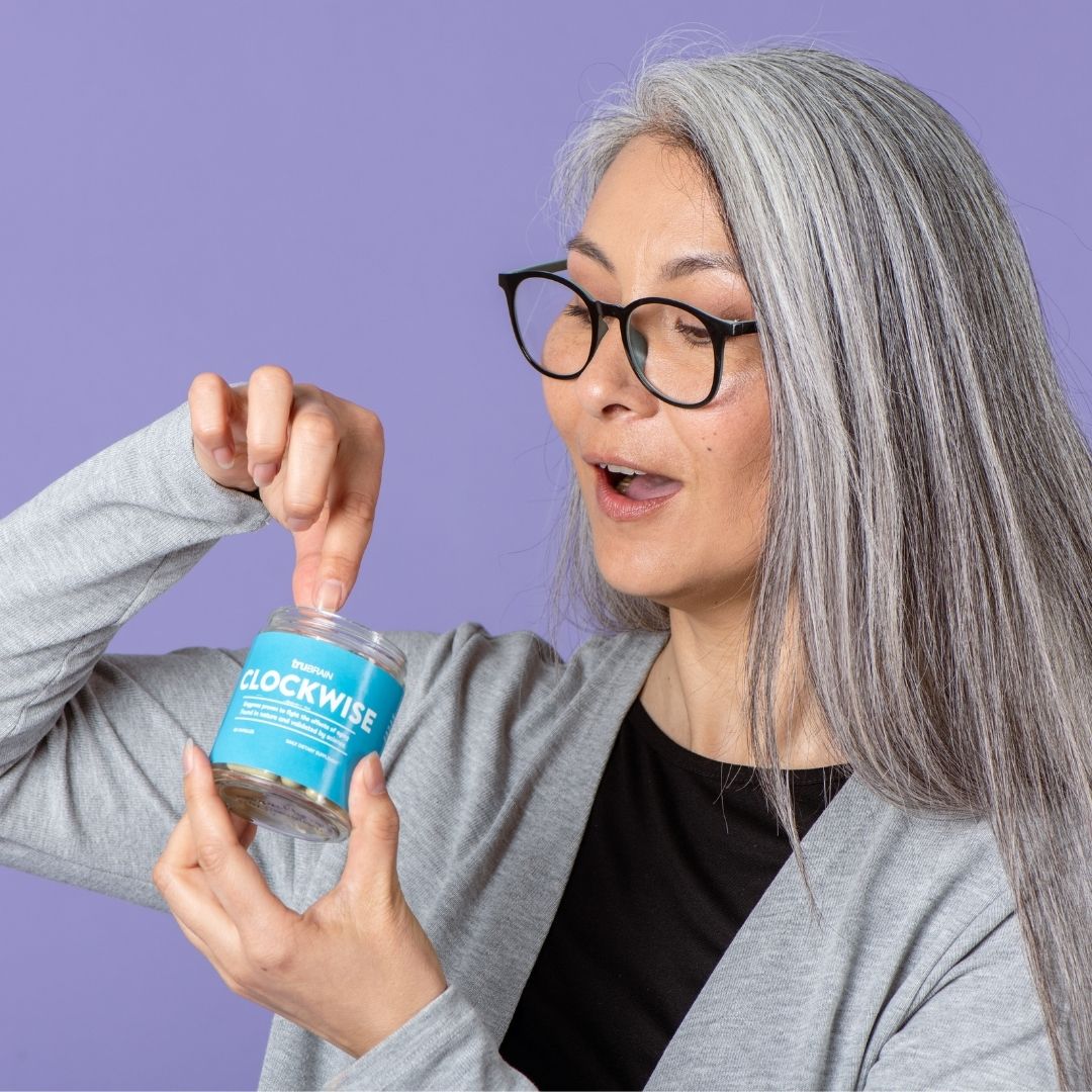 Vibrant 58 year old woman with glamorous healthy, grey hair reaches excitedly into the Clockwise Jar, to take a step on her healthy aging journey.